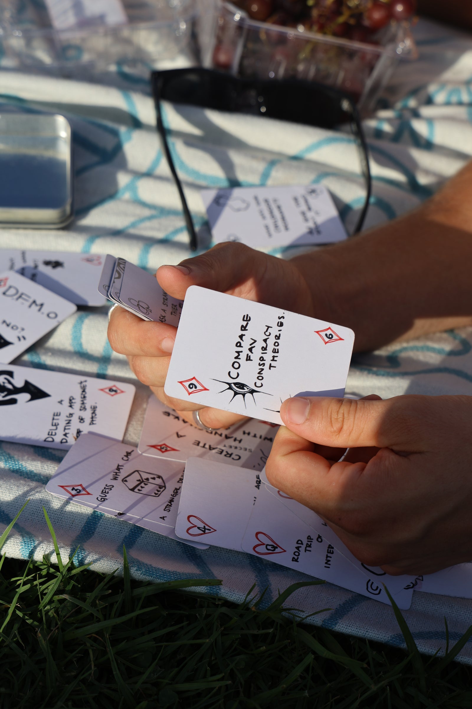 A close-up of a person's hands holding a white playing card that with hand drawn illustration, surrounded by other cards on a blue and white patterned blanket outdoors.