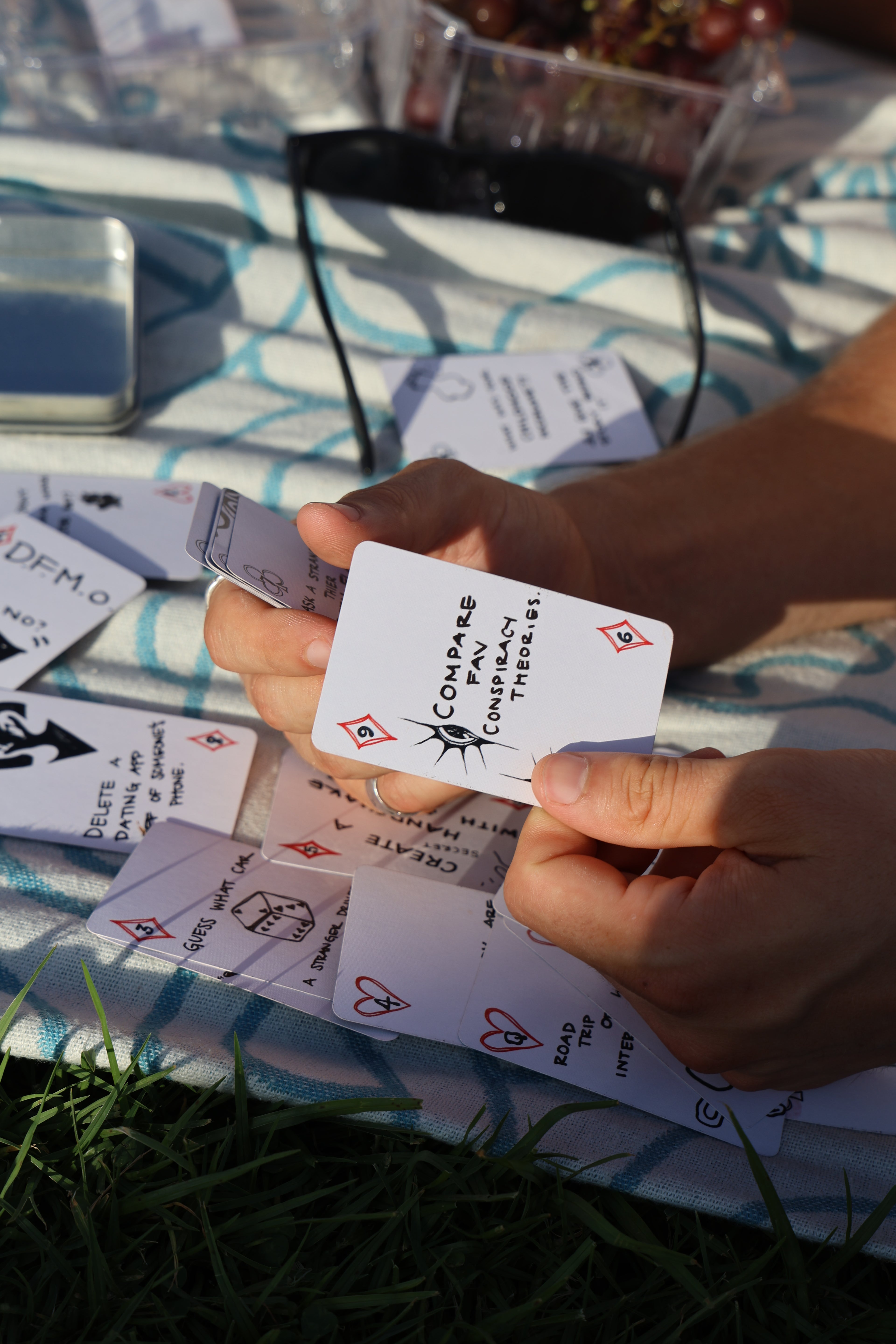 A close-up of a person's hands holding a white playing card that with hand drawn illustration, surrounded by other cards on a blue and white patterned blanket outdoors.